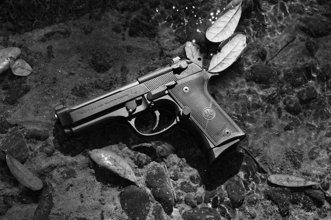 Beretta 92X Compact pistol in a rock bed stream shot in B&W short exposure with leaves floating by on a shallow, crystal clear swath of water about one-inch deep