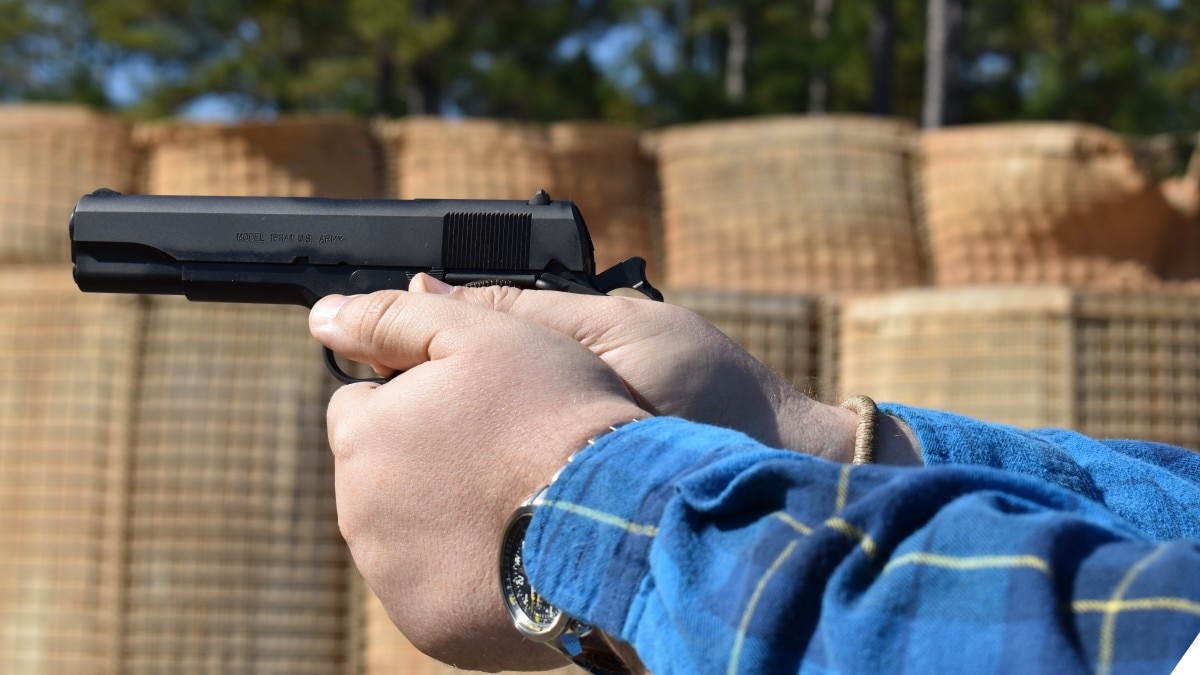 A man in a flannel shirt holding a M1911 pistol at a shooting range