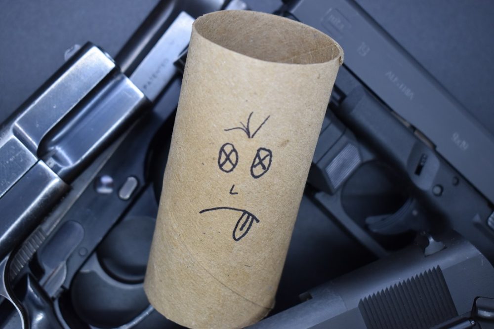 An empty TP tissue roll with a face drawn on it amongst a pile of handguns