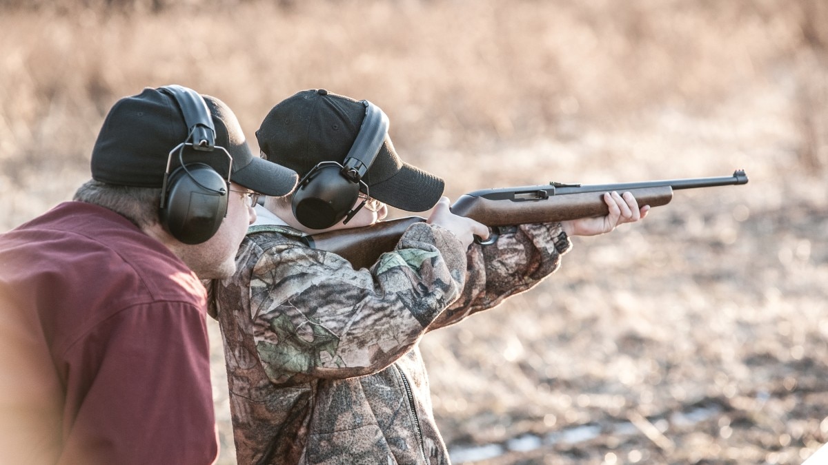 A youth takes aim with a Ruger 10/22 rifle under adult instruction on a shooting range outdoors